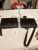 Two black Sylvania portable DVD players side by side on a table with power adapters and carrying case.