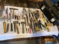 Overview of masonry tools laid on a white sheet including hammers, chisels, trowels, saw, level, pliers, and toolbox.