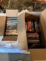 Top view of two open cardboard boxes filled with assorted DVDs and VHS tapes. Boxes rest on a white tablecloth.