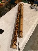 Two rain sticks lying on a white cloth. One is large, with dark burnt wood pattern and woven ends with cord. The other smaller, lighter in color with zigzag line decorations and carved ring details. A hand is visible holding the larger one.
