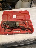 First Milwaukee reciprocating saw inside its red carrying case with attached power cord, shown on white fabric surface.