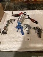 Overview of five paint spray guns and one red and black hand tool arranged on white cloth.