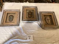 Three framed mask wall art pieces displayed on white cloth, each with a rustic whitewashed frame and mounted mask sculpture.
