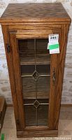 Front view of oak cabinet with glass door and decorative leaded diamond shaped patterns, showing metal handle and MaxSold tag.