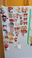 Full view of fridge covered with various Campbell's soup can themed magnets and three Campbell's plush dolls attached to the fridge.