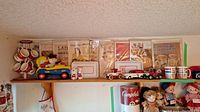 Wide view of shelf with vintage Campbell's items including mugs, model cars, dolls, salt and pepper shakers, apron, and paper advertisements on wall.