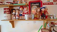Shelf displaying Campbell's Kids dolls and figurines, a metal tub, toy camper van, and other collectible items with Campbell's Soup branding.
