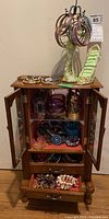 Full view of open wooden jewelry cabinet on wood floor, top surface with bracelets, inside shelves with jewelry, bottom drawer open showing rings