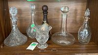 Set of five various crystal and blown glass items including four decanters and one small vase or bowl, arranged on a wooden shelf.