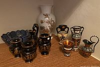 Overview of the lot showing black Greek mini urns with gold trim, white German vase with gold florals, ceramic blue weave bowl, and Greek key pattern mini urns.