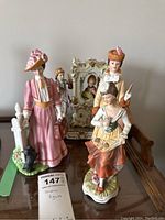 Ceramic figurines arranged in a line including a woman in pink dress, a young woman in yellow apron, and an ornate porcelain panel in the background