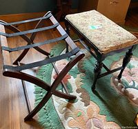 Side and top view of wooden folding luggage stand with black straps and adjacent upholstered stool with floral fabric on seat, both on floral rug over hardwood floor.