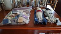 Two vintage model cars next to a plastic bag filled with assorted matchbooks on a wooden surface.