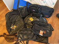 Overview of assorted bags arranged on floor including leather purses, black messenger bags, backpacks, and branded fabric bags