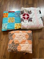 Three folded quilts on floor showing patchwork designs and colors.