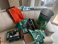 Photo showing collection of Roughriders towels, scarves, license plate brackets in boxes, and a large Saskatchewan Roughriders bucket/container on a white chair.