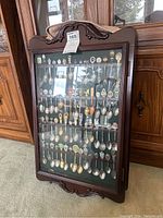 Full view of wooden dark-finish display cabinet holding multiple rows of souvenir spoons with glass front closed