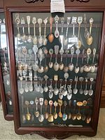 Full view of the display cabinet with approximately 50 souvenir collector's spoons hung inside on multiple horizontal rails against a dark green background.