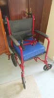 Front view of wheelchair showing red frame, blue padded seat, black backrest, and armrests with padding.