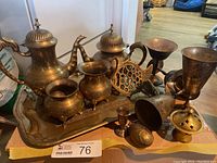 Full view of all brass and silver plate items in the lot on a low surface with wood floor background.