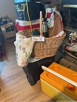 Wicker picnic basket with floral cloth, metal thermos, green kettle, and other picnic items visible inside and around the basket on a rolling cart.