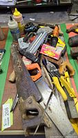 Various carpentry hand tools including hand saws, pliers, staple gun, hammer, measuring tape, and rusty tools on a work bench