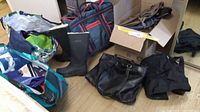 Shows multiple colorful cloth grocery bags, a black duffle bag with red details, a pair of black Tretorn lined rubber boots size 38, and a large black leather handbag and some other black bags on a wooden floor.
