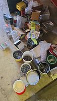 Wide shot of mixed hardware containers on floor including tubs, tins, boxes with screws, nails, bolts, and nuts.