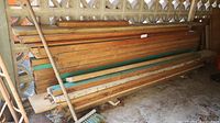 Assorted lumber boards stacked horizontally against wall beneath shelter; includes 2x4s and wider flat boards, some with green paint on ends.
