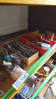 Photo showing a box full of assorted metal wrenches including adjustable and fixed sizes, screwdrivers and a metal tiger torch on a shelf.