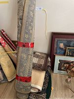 Stack and rolled large area rug with floral patterns tied with red bands among other rugs and some framed art.