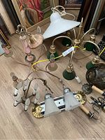 Wide view of multiple vintage light fixtures including pink brass chandeliers, white pendant light, and green glass and brass fixtures arranged on floor.