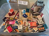 Large clear plastic container filled with assorted vintage matchbooks and two ashtrays.