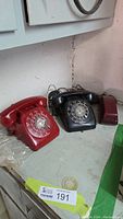 Photo showing two rotary dial phones side by side: one red phone in good condition, and one black phone with handset and wiring visible.