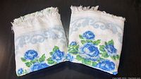 Two folded vintage white bath towels with blue rose floral designs and fringed edges on a black surface.