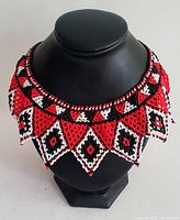 Full view of the handcrafted beaded collar necklace draped on a black mannequin bust, showing the vibrant red, white, and black color pattern and collar shape.