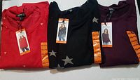 Three folded long sleeve knit tops in red, black with silver stars, and purple, with tags attached.