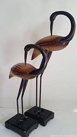 Front view of two wooden and metal stork sculptures on black square plinth bases, showing full height and shape.
