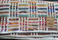 Close-up of folded rug showing multicolored woven pattern and woven white edges, with Rivancia label stating 'Hand Woven Rug, Made with Care'