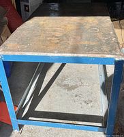 Front-side angle view showcasing the blue steel frame and heavily worn metal tabletop with discoloration and marks.