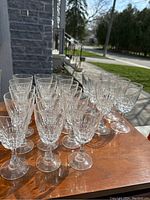 Wide shot of 20 crystal wine glasses arranged on a wooden surface in sunlight, showing both shorter and taller glasses in groups.