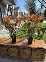 Two tall artificial wheat grass planters on a wooden chest outside in daylight.