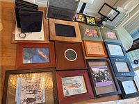 Top down view of assorted picture frames arranged on a wooden floor and black suede photo albums stacked on the left side, showing a variety of sizes and shapes.