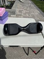 Front view of the black hoverboard placed on a white table outdoors, showing footpads and wheels.
