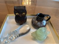 Top-down photo showing all four items: wood owl, pottery pitcher, glass hippopotamus paperweight, and jade dog carving on white tray