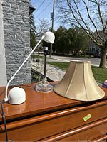 Photo of the three lamps on a wooden surface outside showing a modern adjustable white and clear desk lamp, a clear table lamp base, and a camel colored shade.