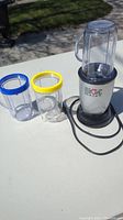 Magic Bullet blender motor base with three clear plastic canisters with colored rims shown together on a white table outdoors.