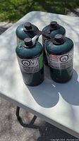 Four green Coleman propane gas canisters arranged in a square on a white table outdoors.