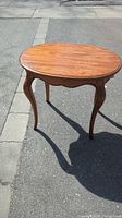 Full view of the oval oak side table showing scalloped edge apron and cabriole legs, placed outdoors on pavement.