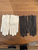 Two pairs of gloves laid flat on wood surface, one black pair with bow detail and one ivory pair with scalloped edge.